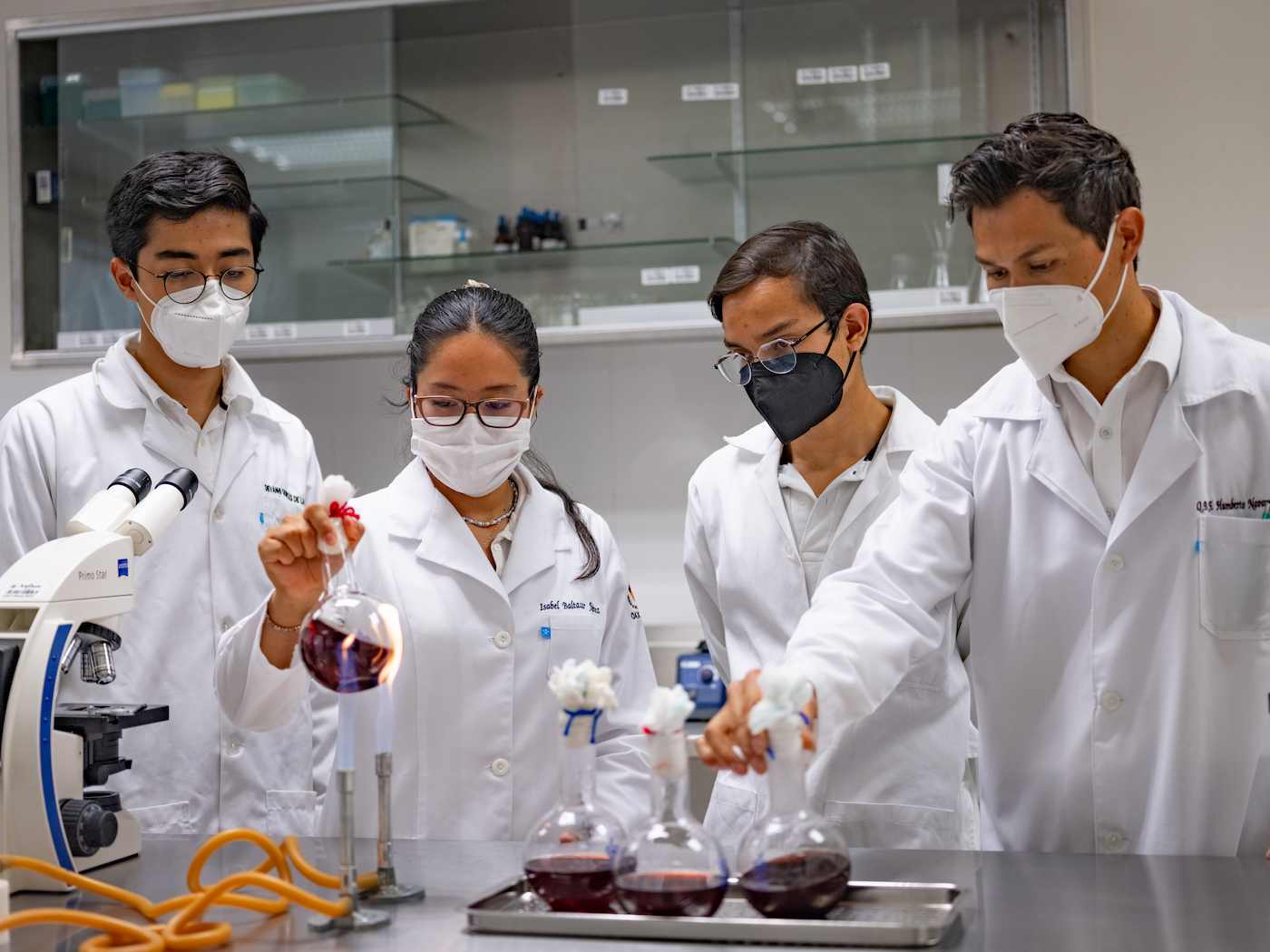 Cuatro personas con bata y mascarilla realizan un experimento con matraces y líquidos color vino en un laboratorio químico.